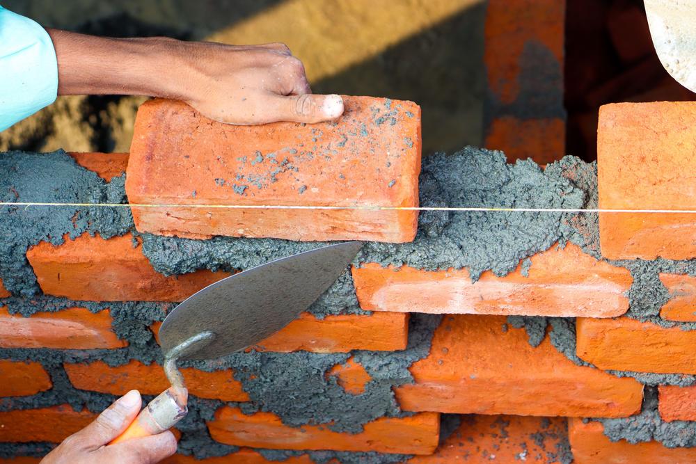 Close-up of a bricklayer spreading mortar with a trowel and laying red bricks to a string line.