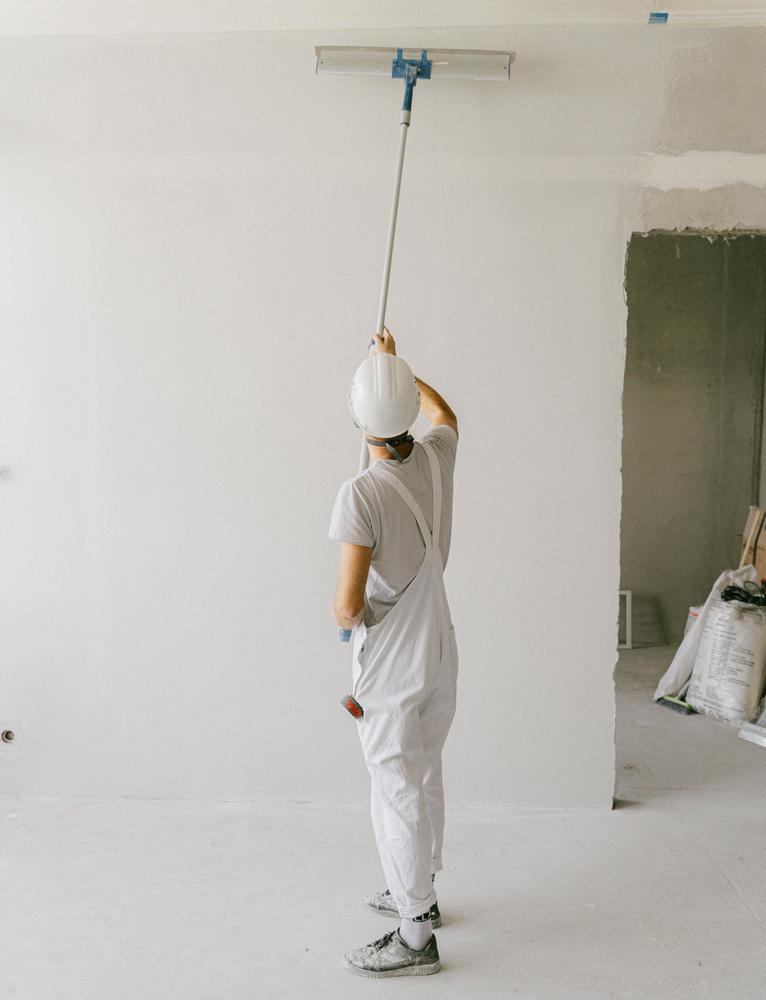 A decorator in white overalls and a hard hat painting a ceiling with a long-handled roller in an empty room.