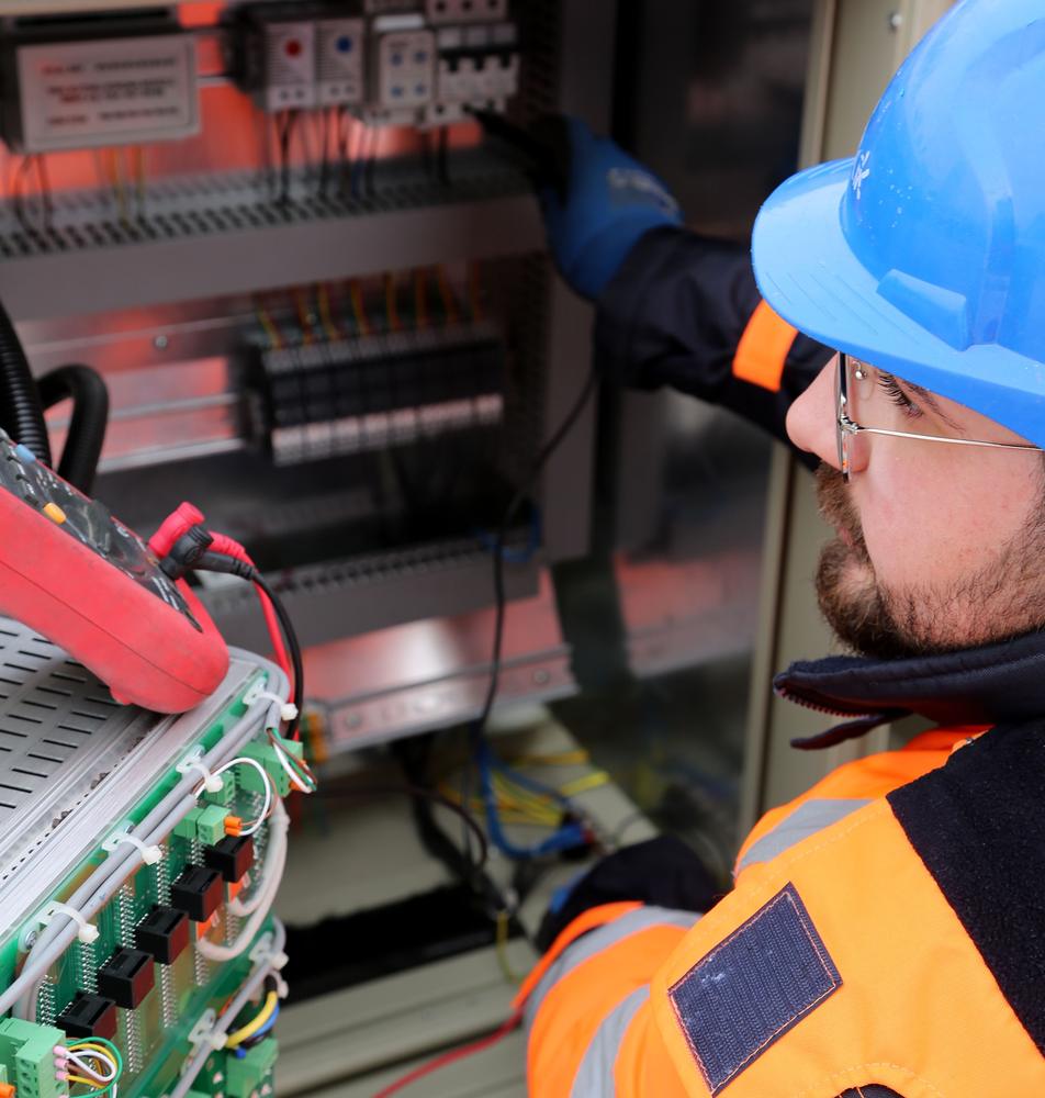 An electrician in a blue hard hat, safety glasses and orange hi-vis jacket testing wiring inside an electrical control cabinet.