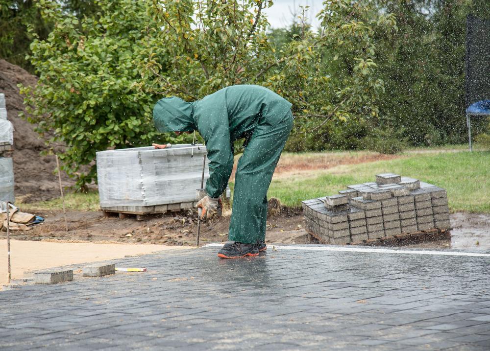 A worker in waterproof overalls laying block paving on a wet day, with pallets of paving blocks nearby.